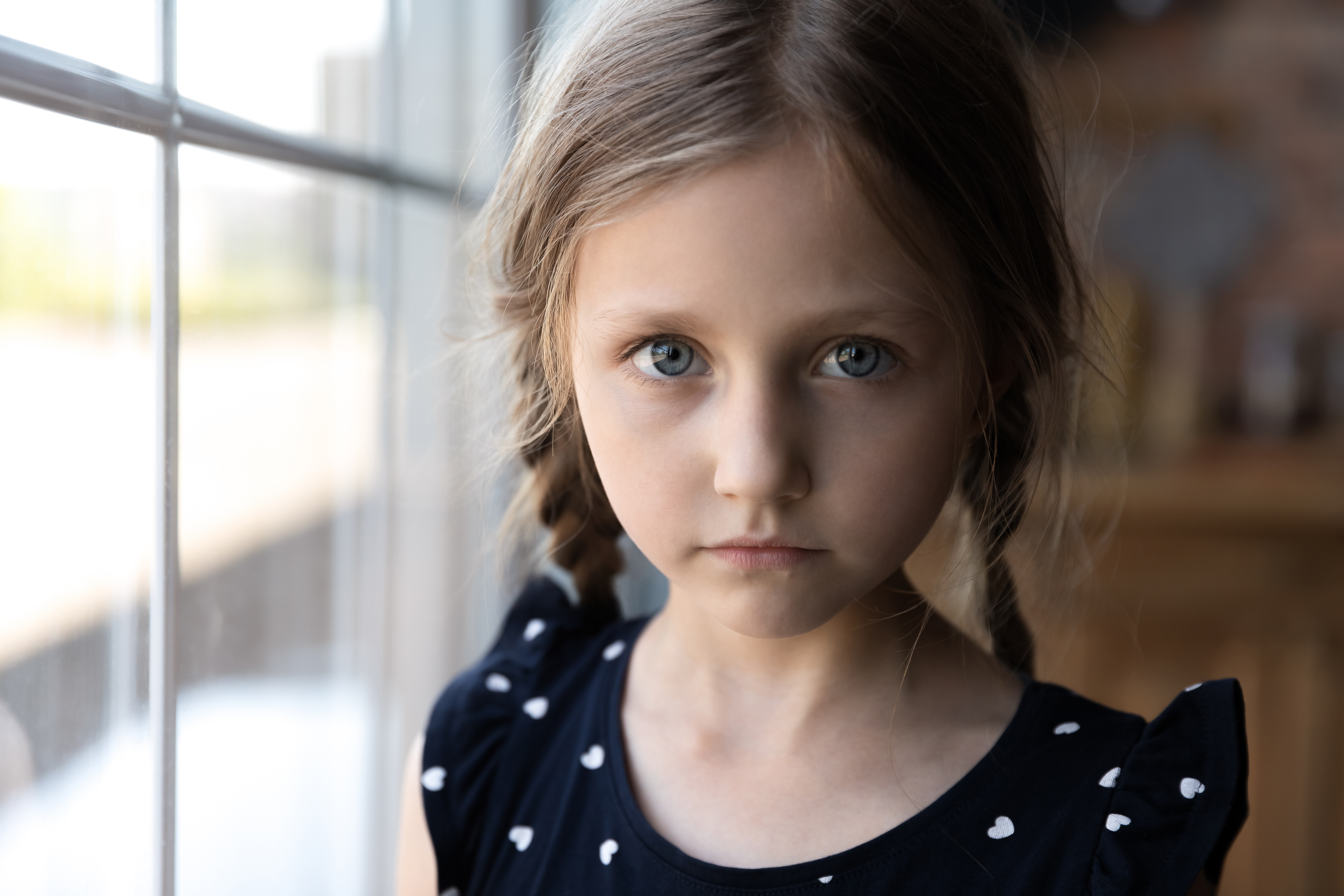 Close up portrait of pensive serious little school preschool age girl standing by window at home looking at camera with sad unhappy face having problem trouble thinking about bad relations in family